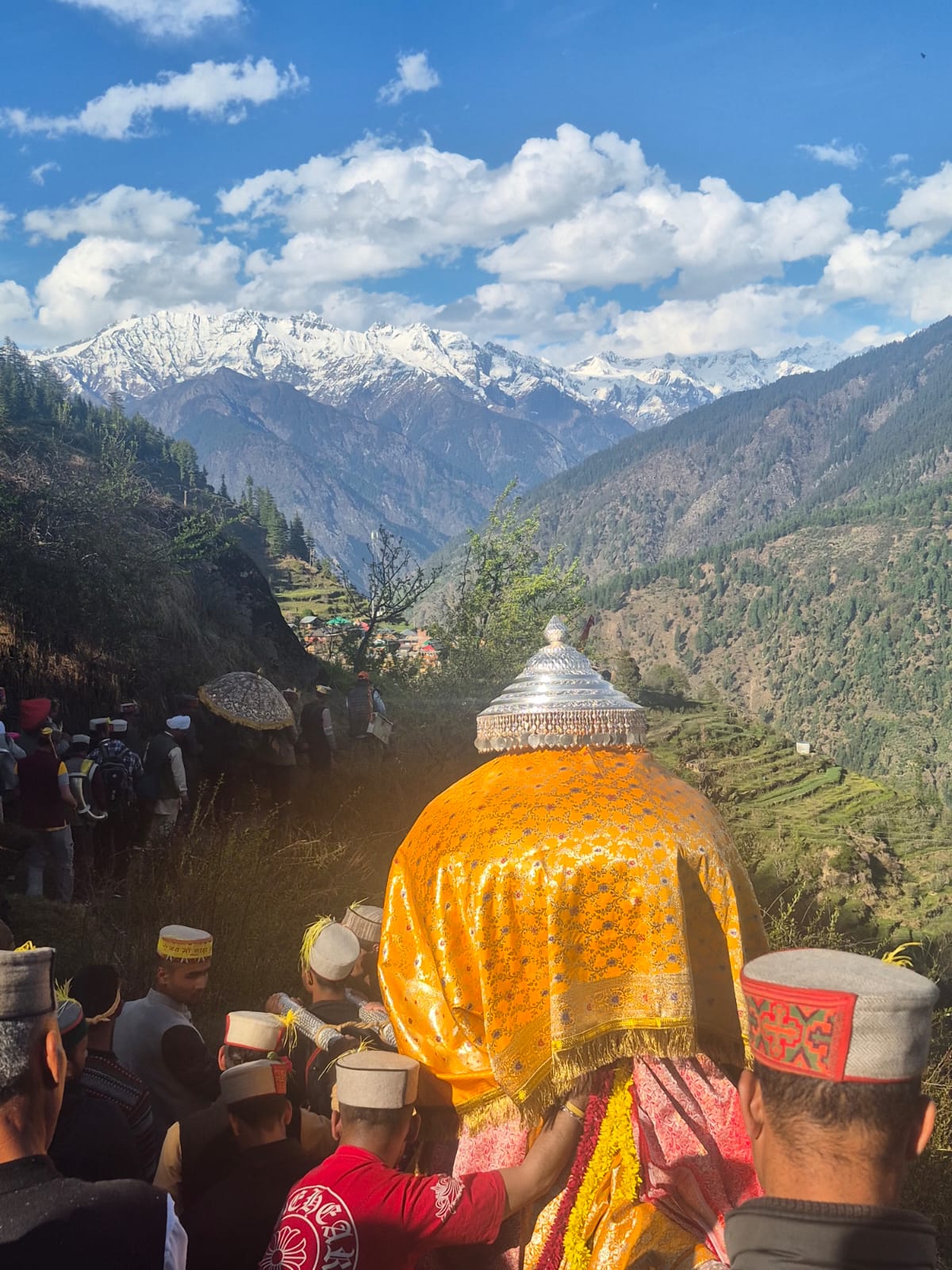 A village devta procession moving through a Himalayan valley beneath snowy peaks