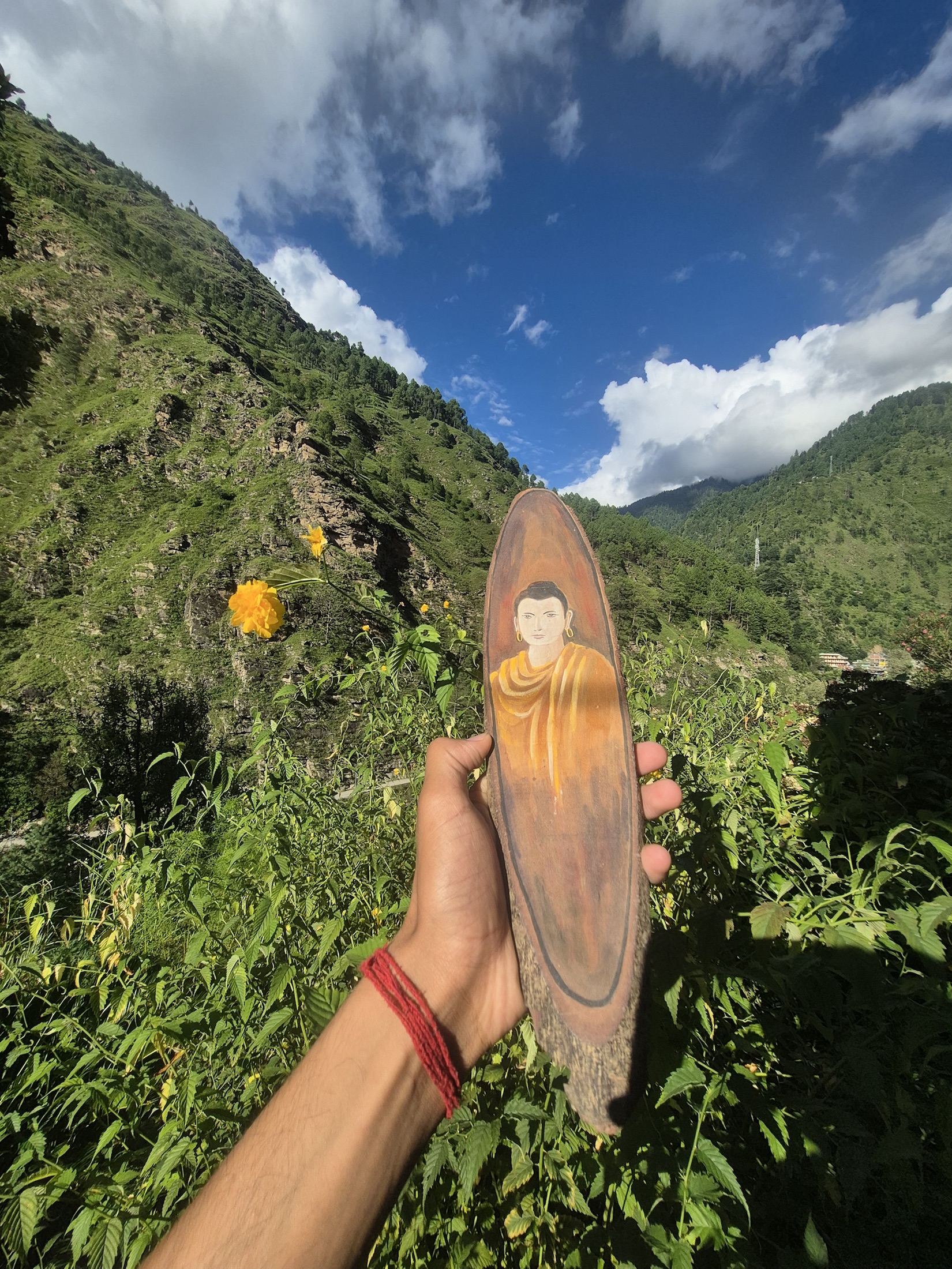 A hand holding a Buddha portrait painted on wood against green Himalayan slopes